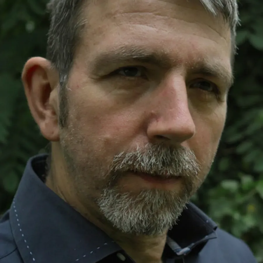 Portrait of the sculptor Ralf Kleine with a greying beard in front of green plants