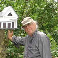 Sculptor Raimund Schmelter wearing a cap in the garden next to a white birdhouse shaped like a villa